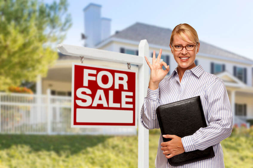 A person with a striped shirt and glasses smiles, holding a black folder and showing an OK gesture next to a "For Sale" sign in front of a house.