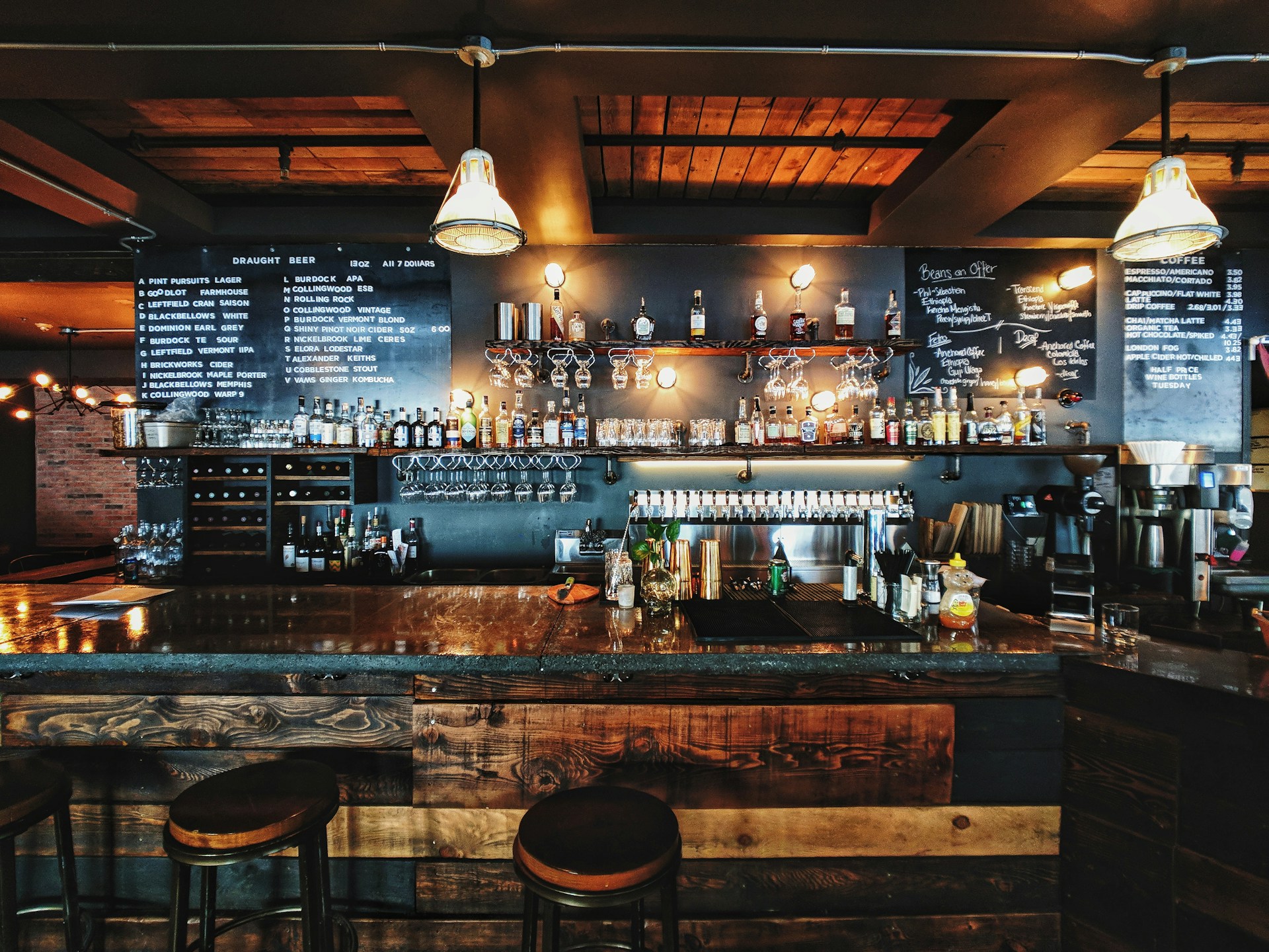 A well-lit bar with wooden counters, stools, and shelves of liquor bottles. A chalkboard menu hangs above the bar listing drinks. Glasses are hanging upside down. Two overhead lights illuminate the area.