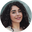 Woman with curly dark hair and light skin smiling slightly at the camera. She is wearing a white top and is positioned against a neutral background.