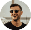 A man wearing dark sunglasses and a black shirt is smiling outdoors.