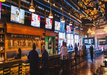 Patrons sit at a long bar with brick and wood decor under exposed industrial ceilings, watching multiple TVs. The bar is well-lit with hanging lights, and a few people are seen standing or sitting.