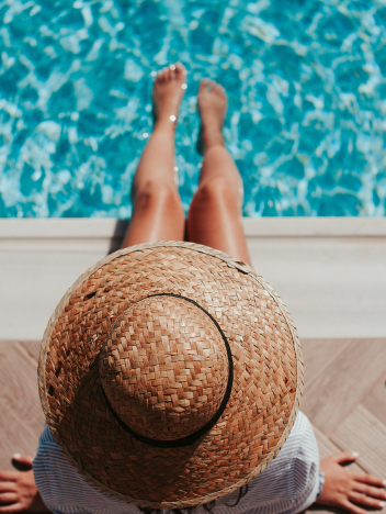 Person wearing a straw hat sitting at the edge of a pool with their feet dipped in the water.
