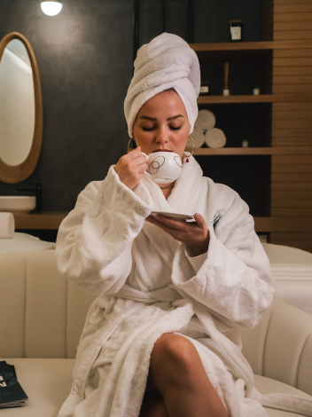 A person in a white bathrobe and towel headwrap sits on a sofa, holding a teacup and saucer, sipping tea in a dimly lit room with shelves in the background.